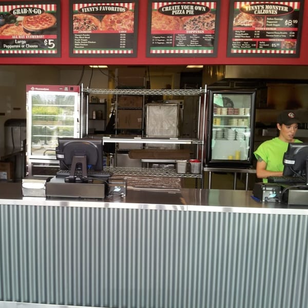 A pizza restaurant counter with menu boards, a refrigerator, and an employee working at a computer.