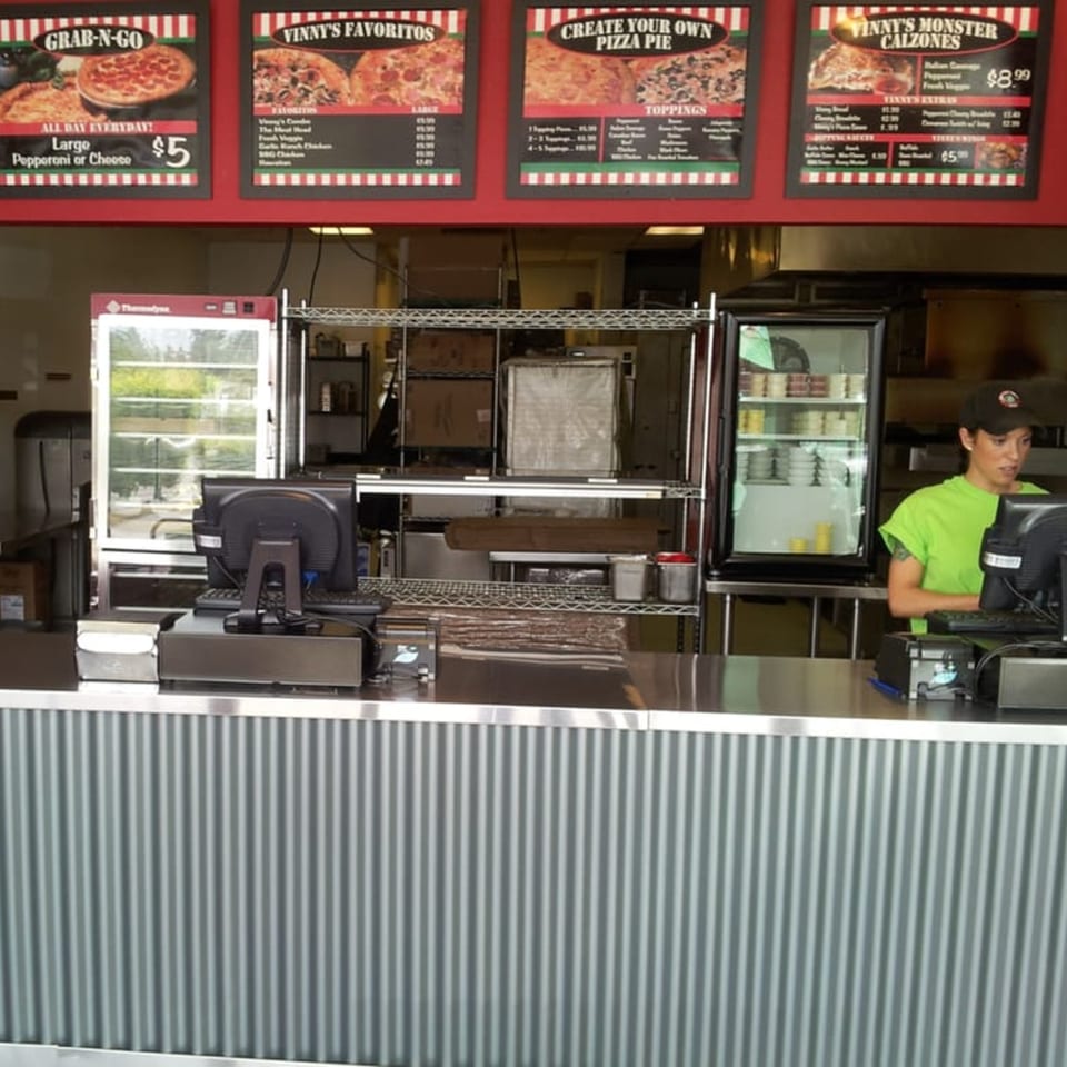 A pizza restaurant counter with menu boards, a refrigerator, and an employee working at a computer.