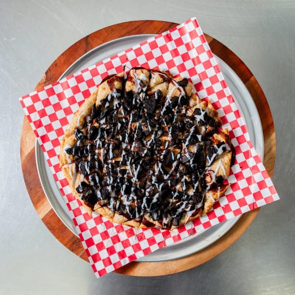 New York Vinny's Pizza dessert pizza with crushed Oreos and white icing drizzle on checkered paper.