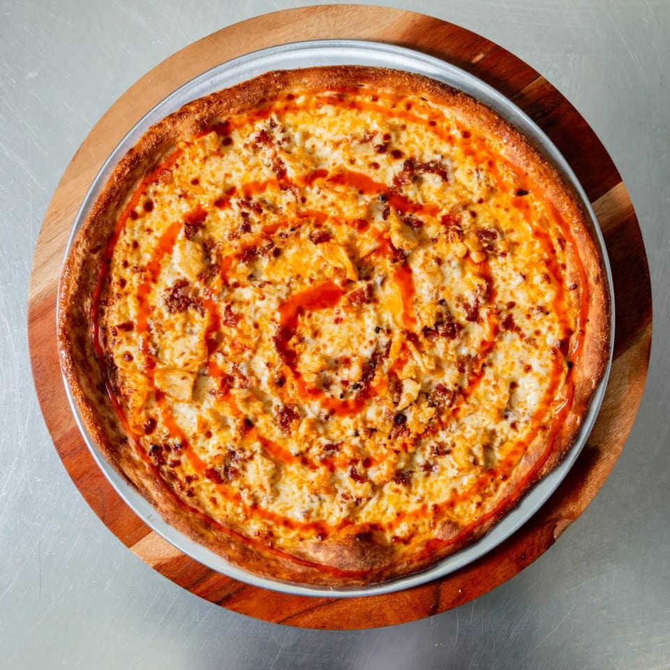 New York Vinny's Pizza's buffalo chicken pizza, drizzled with hot sauce, on a wooden board.